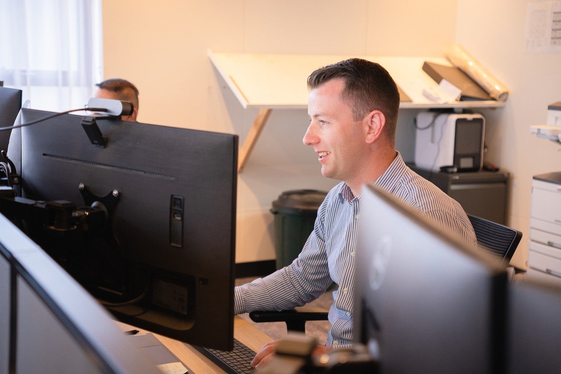 Man in striped shirt sitting and smiling at a computer monitor in an office setting.