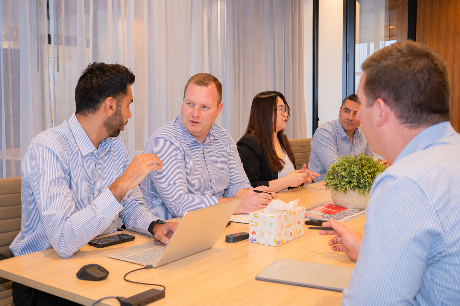 Five professionals having a meeting around a table with laptops, documents, and a potted plant.