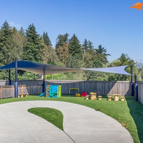 Outdoor playground with shade sails, small climbing structure, and circular concrete path on grass.