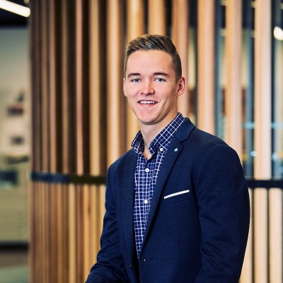 Young man smiling, wearing a navy blazer and checked shirt, with wooden panel background.