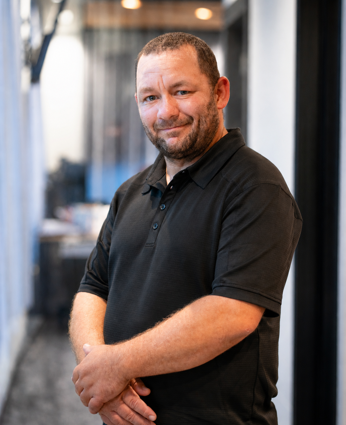 Smiling man in black polo shirt standing with arms crossed in a softly lit hallway.