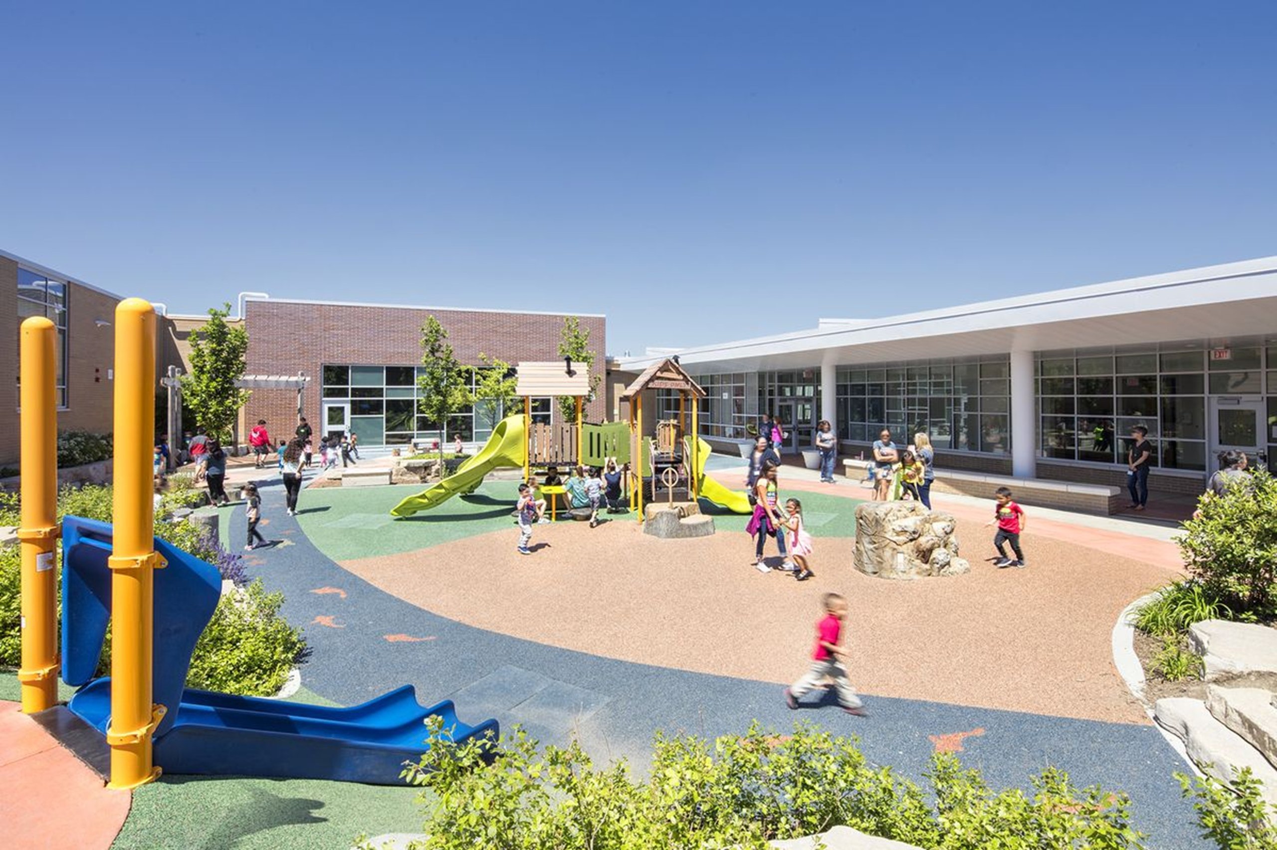 Children playing on playground with slides and climbing structures outside a school building.
