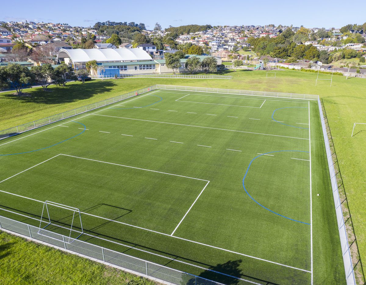 Aerial view of an empty green artificial turf sports field with white and blue markings and fenced edges.