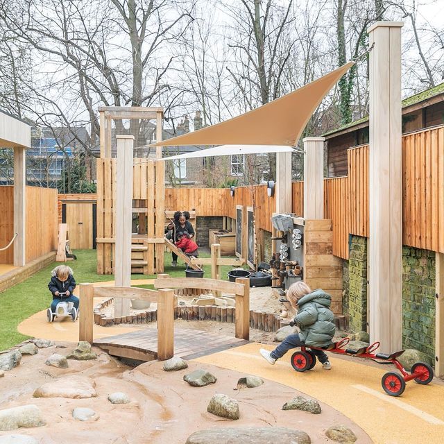 Children playing in a wooden playground area with a bridge, climbing structures, and ride-on toys.