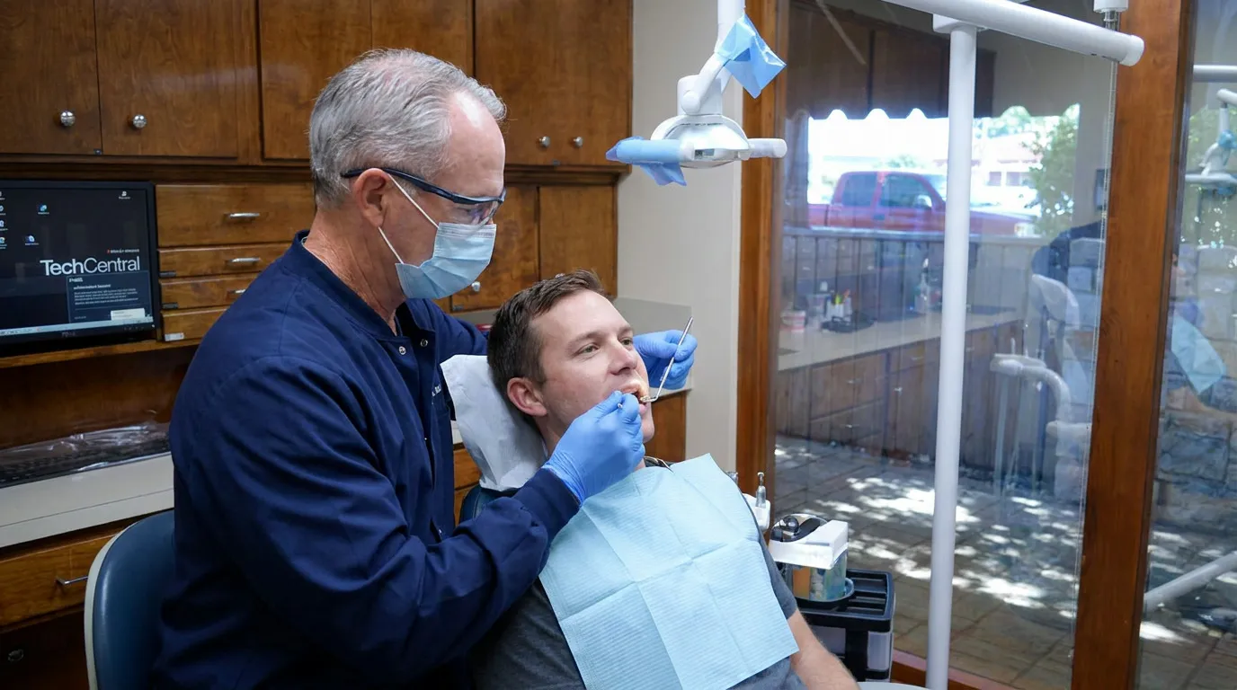 A dentist in blue scrubs, mask, and glasses examines a patient in a dental chair with a mirror and probe, while a computer monitor and wooden cabinets are visible in the background.