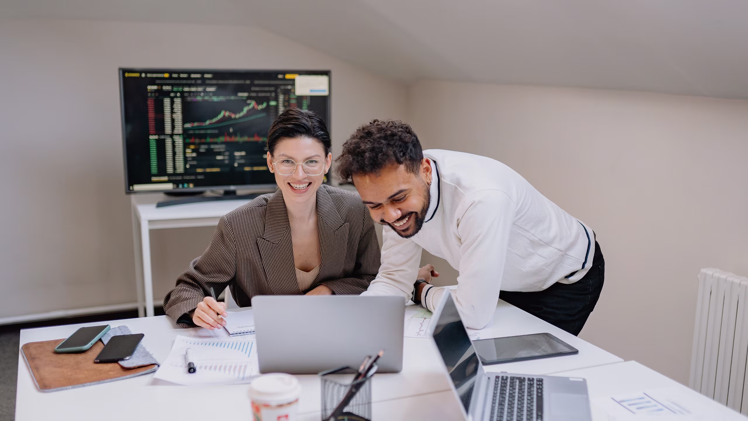 Two coworkers smiling and working together on laptops with charts and phones on the table.