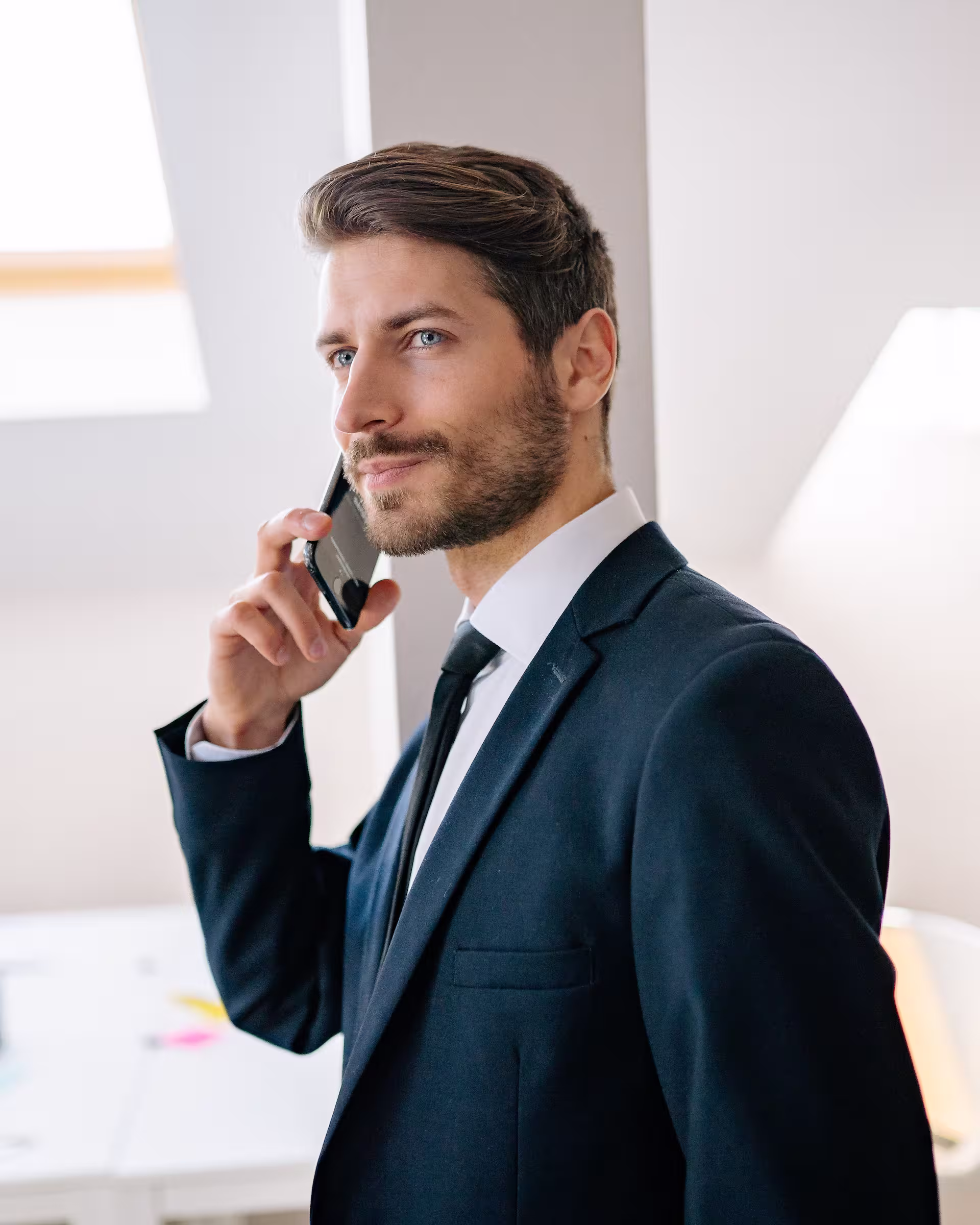 Man in a black suit talking on a smartphone in a bright office space.