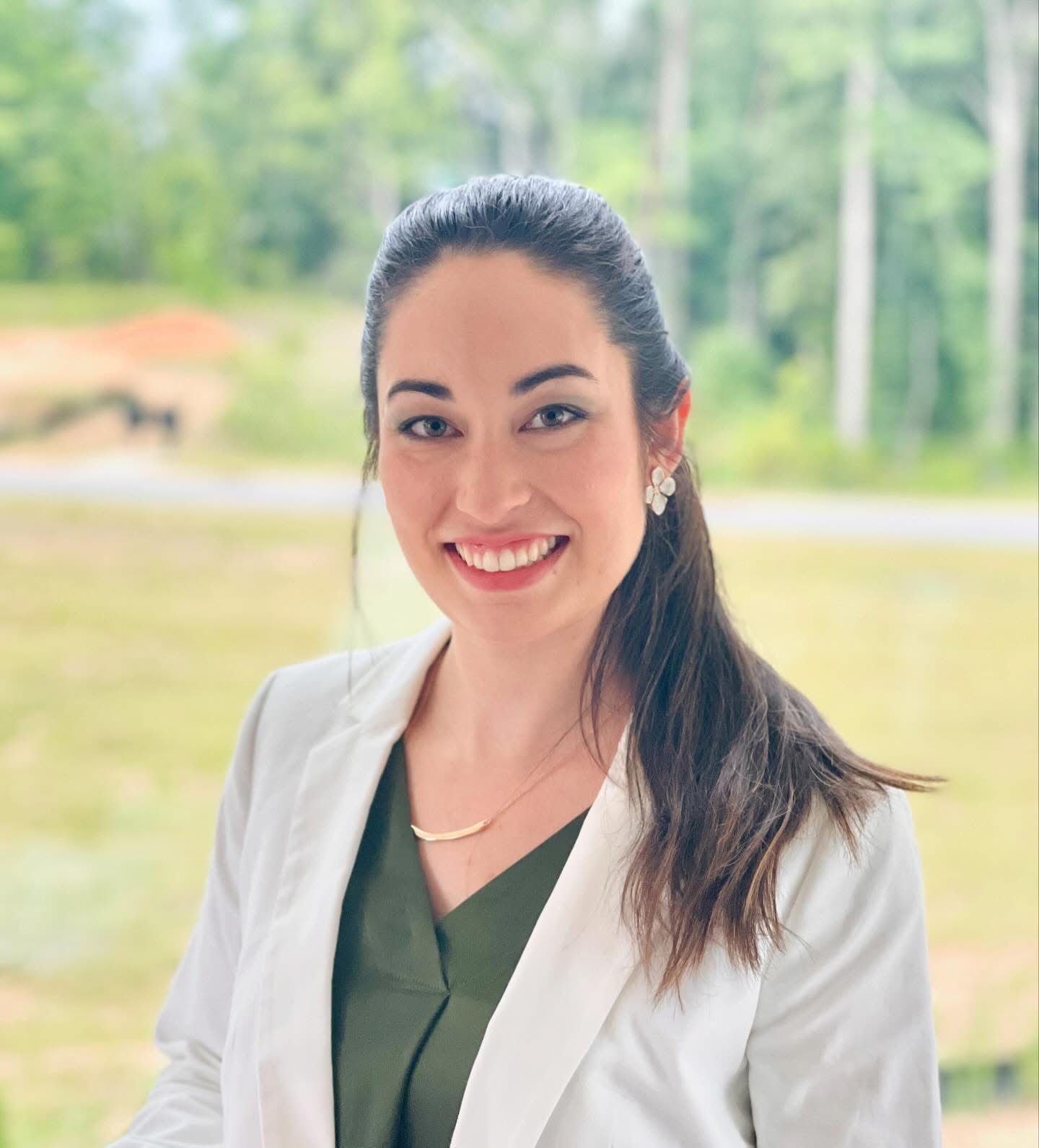 Smiling woman with long dark hair wearing a white blazer and green top outdoors.