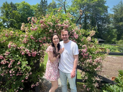 Young couple smiling and posing in front of a wooden arch covered with pink flowering vines.