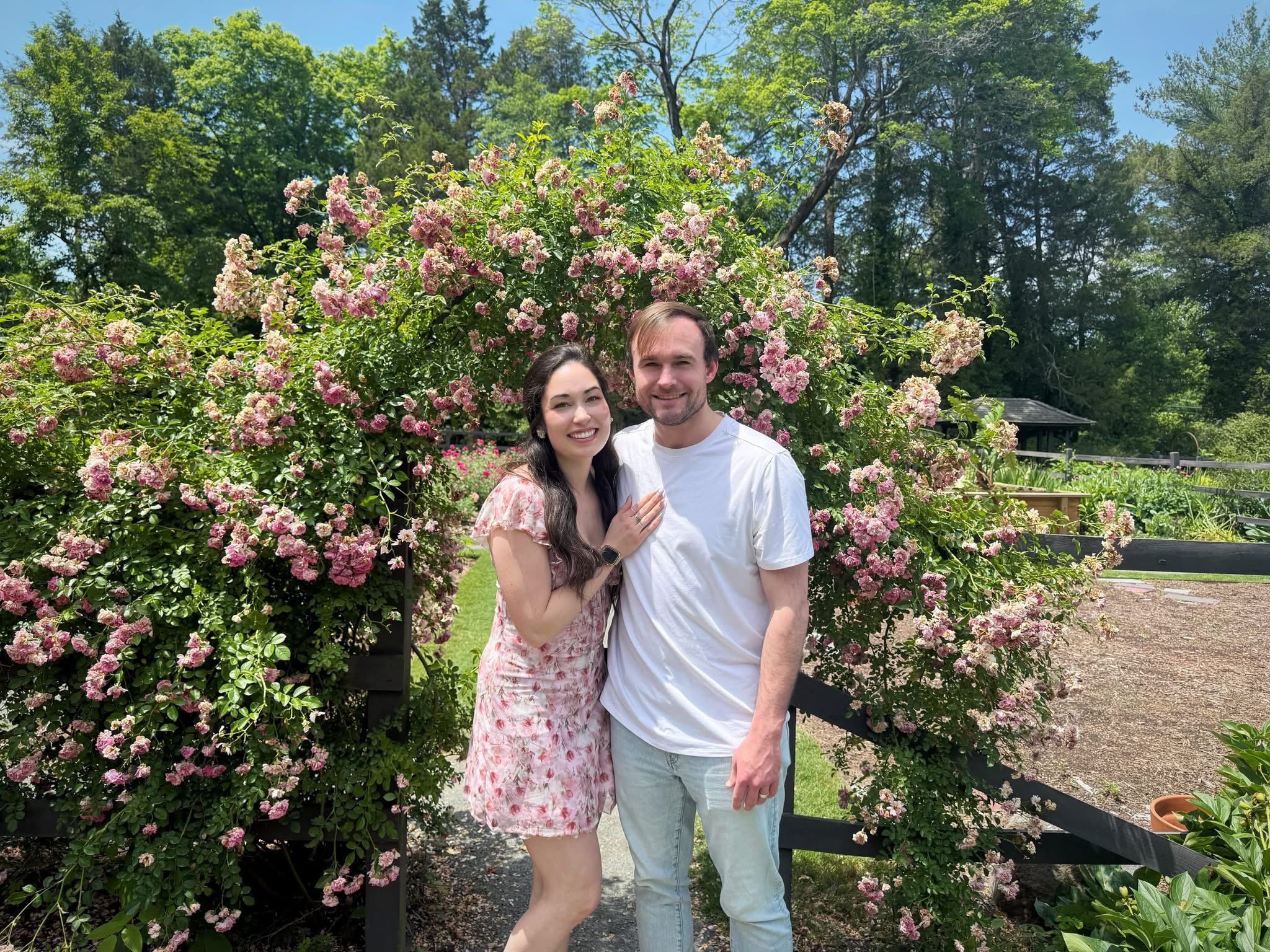 Young couple smiling and posing in front of a wooden arch covered with pink flowering vines.