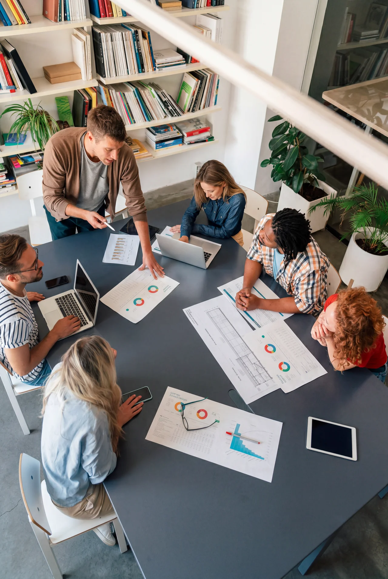 Six people collaborating around a table with charts, laptops, and papers in a bright office space.