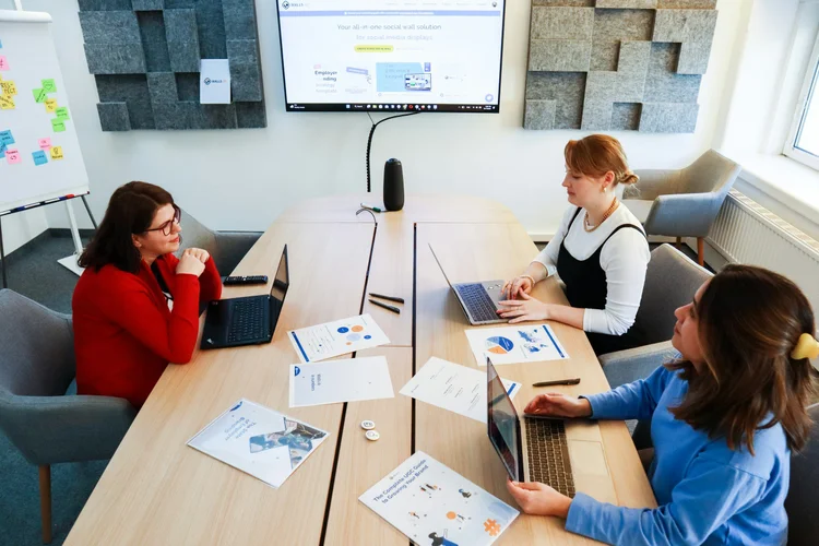 Three women working with laptops and papers around a conference table in a modern office meeting.
