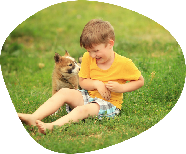 A laughing boy sits on the grass with a small tan and white puppy beside him.