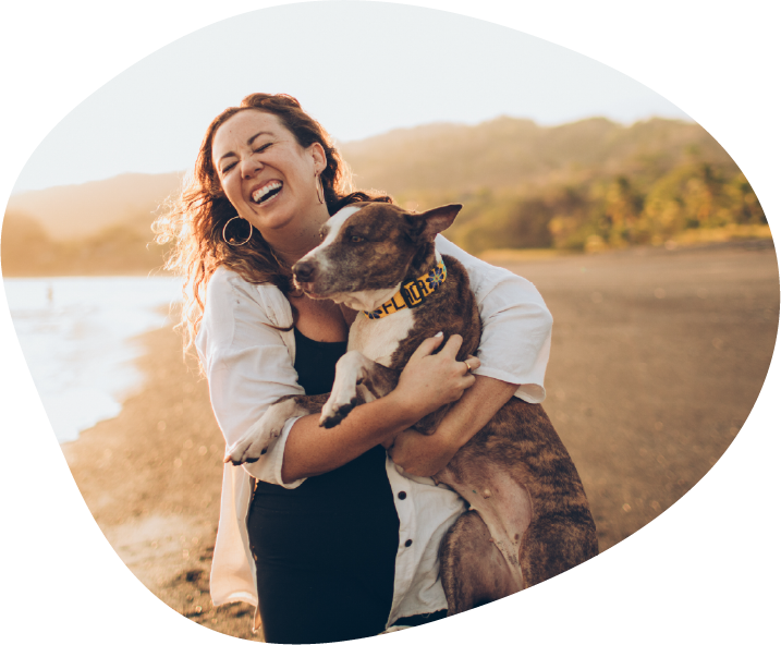 A woman laughs as she holds her brindle dog in her arms on a beach at sunset.