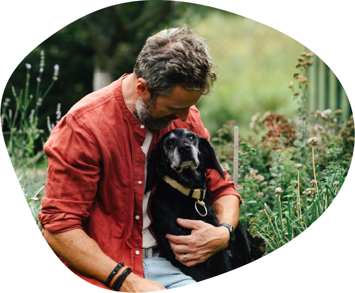 A man kneels in a garden, hugging his greying black dog.