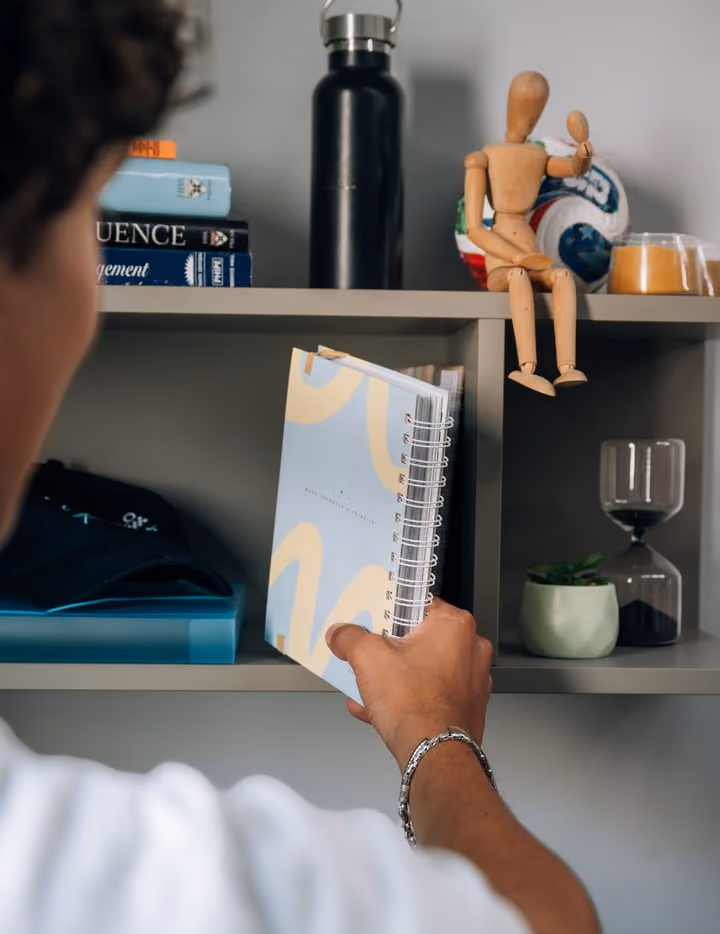 A hand writing in a notebook on a shelf above a desk