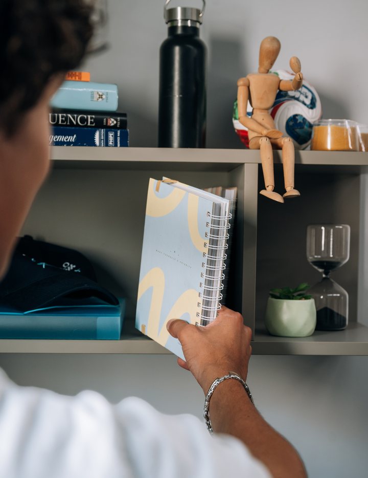 A hand writing in a notebook on a shelf above a desk