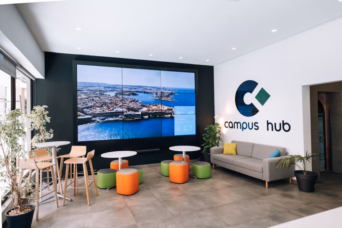 The Campus Hub reception, with a video wall showing an aerial view of Valletta, café-height tables and stools, and the Campus Hub logo on the wall