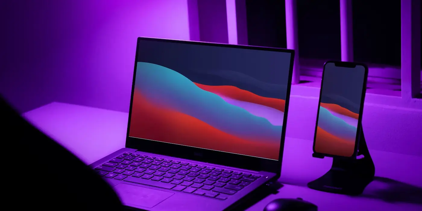 Laptop and smartphone with matching colorful wave wallpapers on a desk lit by purple light.