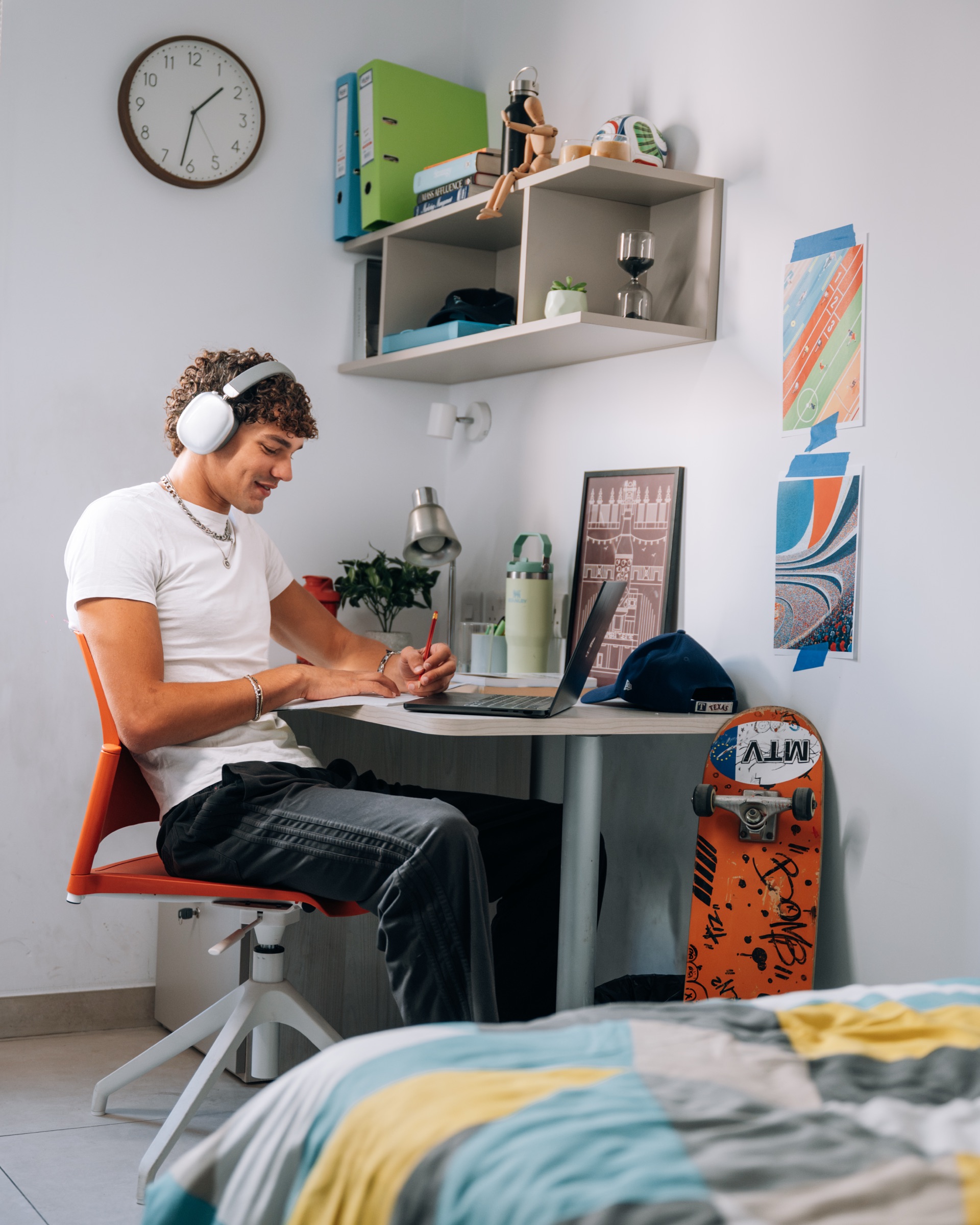 Student in headphones writing at the desk of a Cosy room, laptop open and a skateboard propped against the wall