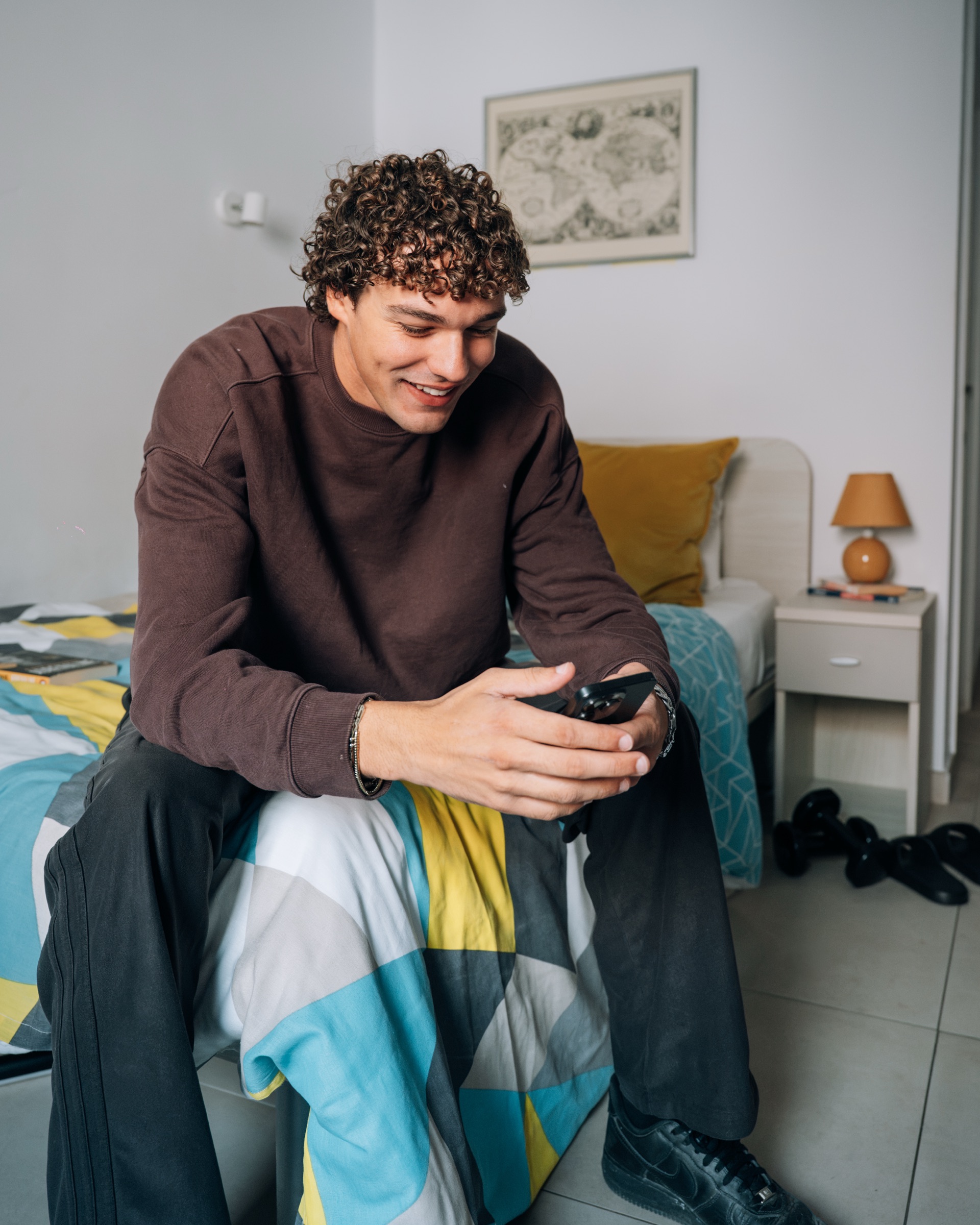 Student sitting on the end of the bed in a Cosy room, smiling at his phone