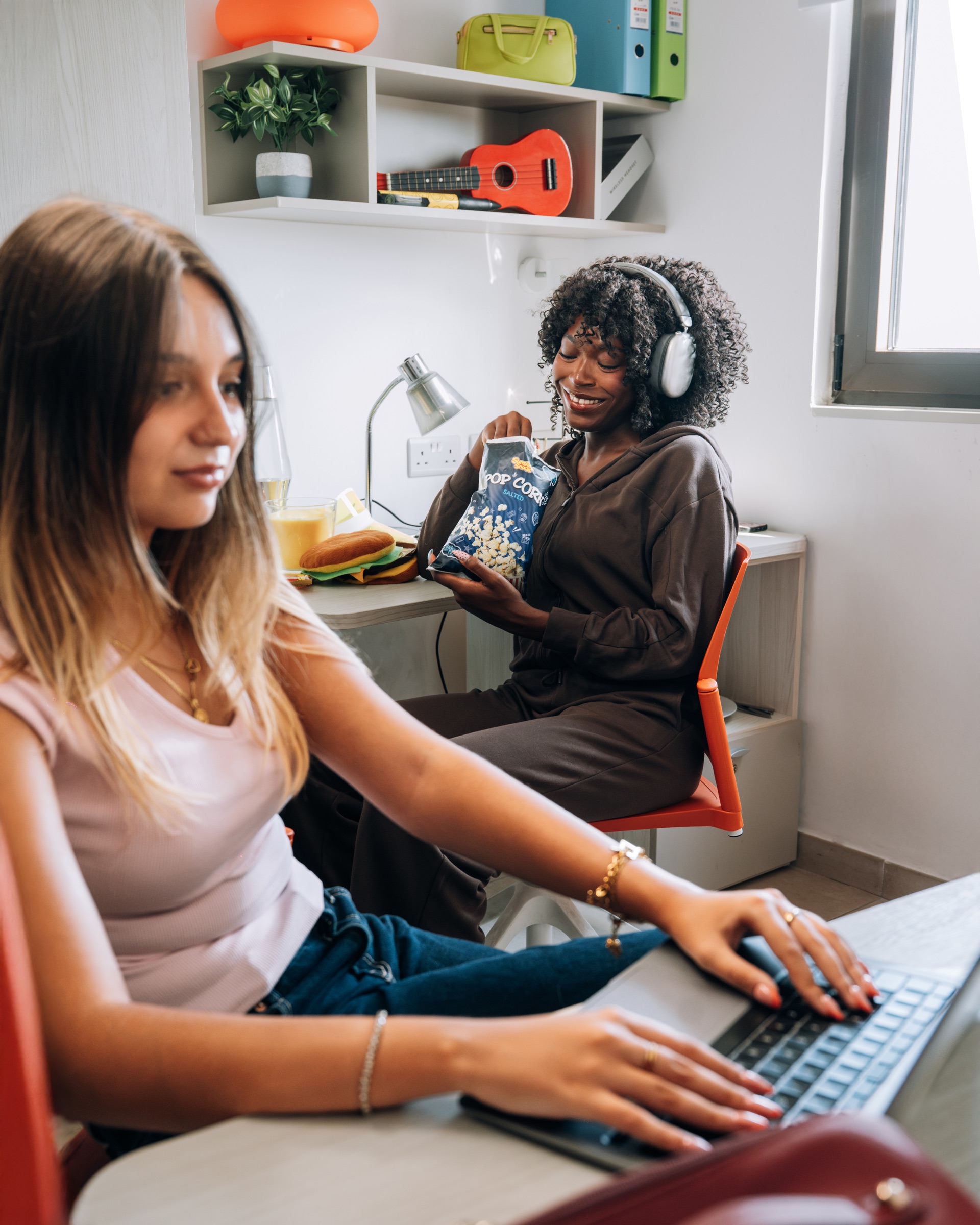 Two students in a Social room, one typing at a laptop while the other shares a bag of popcorn