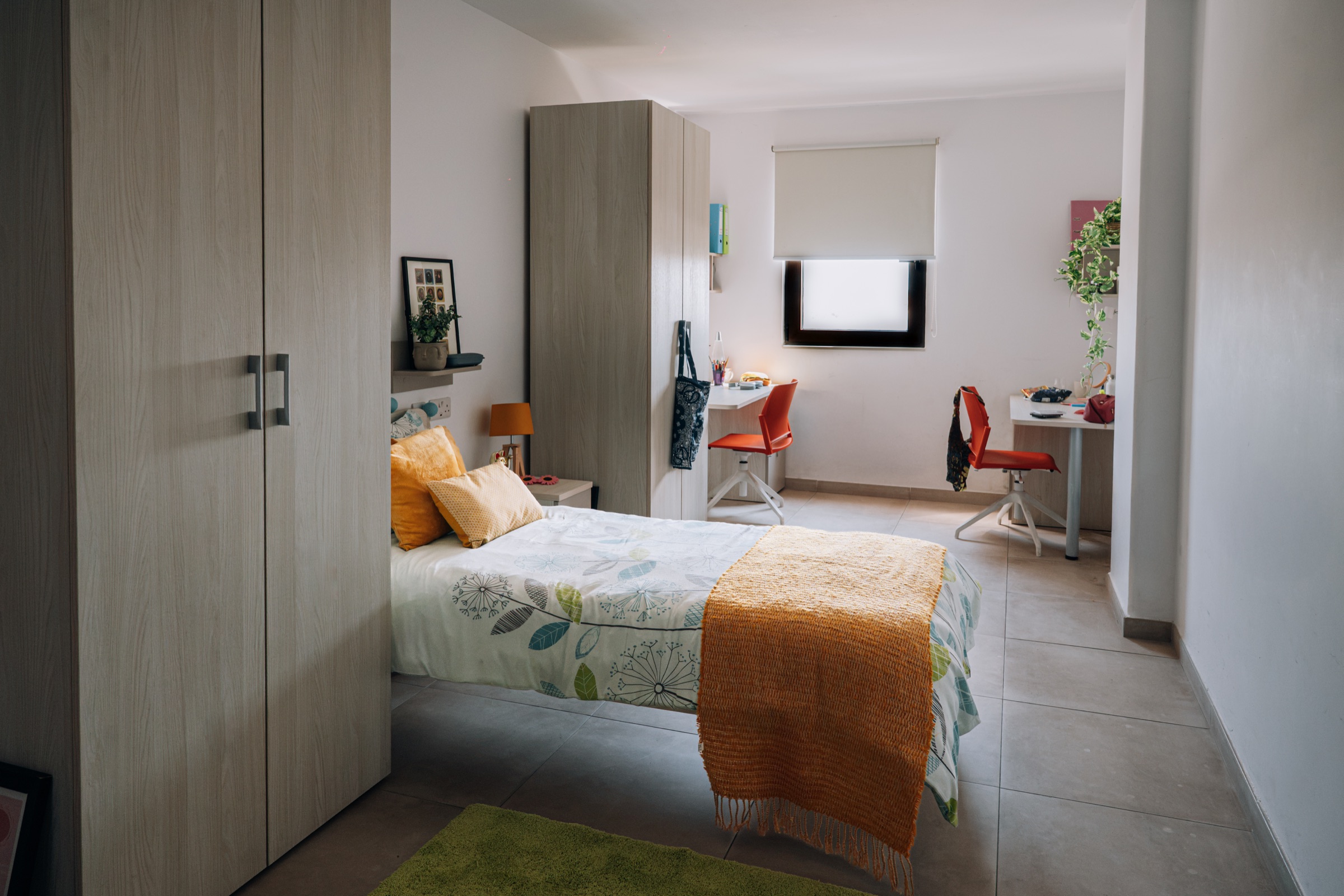 Social room with a single bed and wardrobe in the foreground and two desks by the window