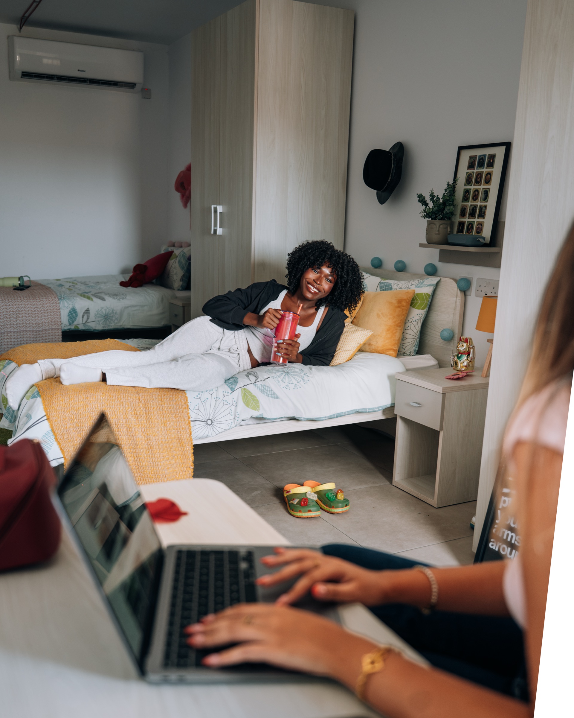 Student relaxing on her bed with a drink while her roommate works at a laptop in the foreground