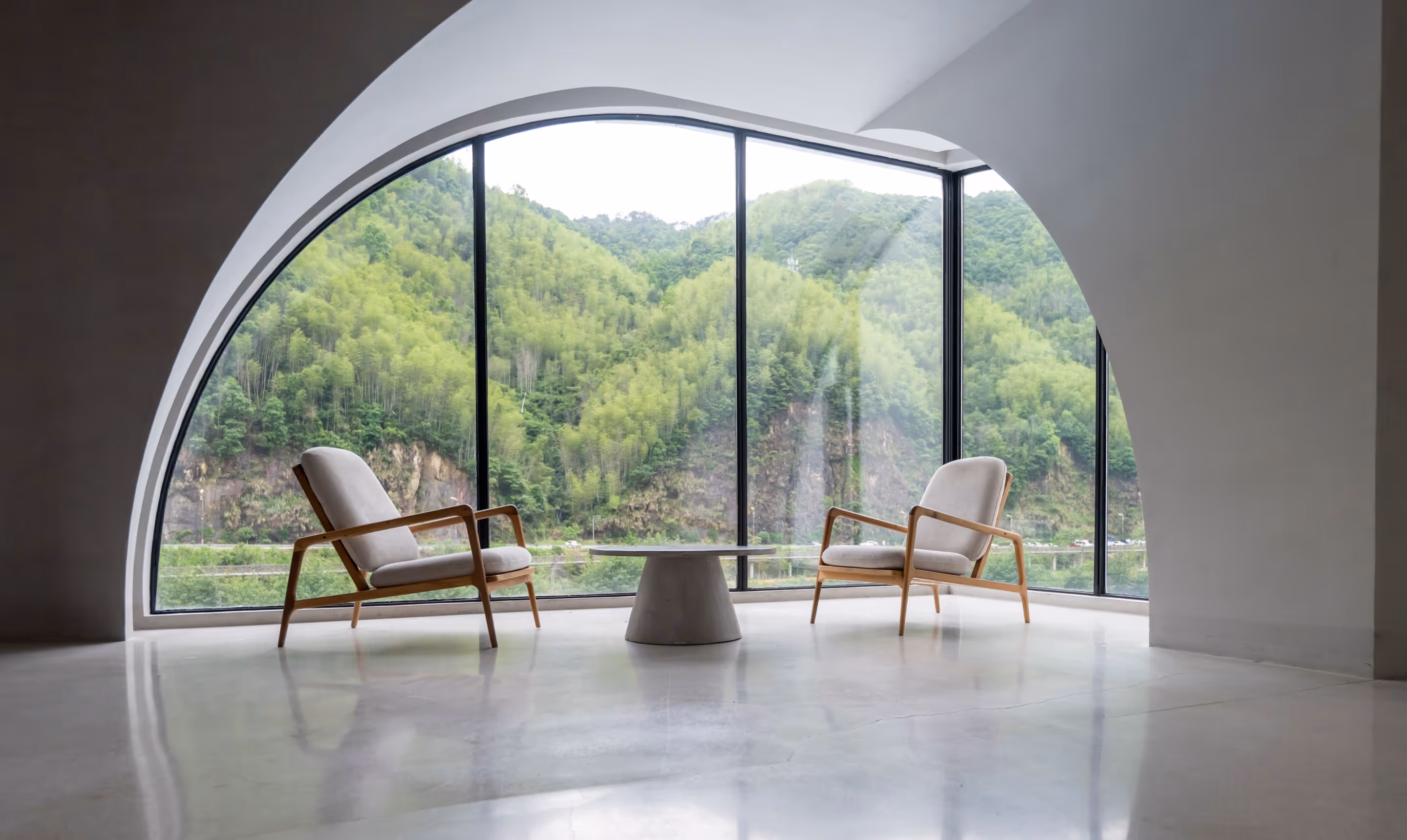 Heller, moderner Raum mit großem Bogenfenster und Blick auf bewaldete Berge, zwei Sessel und ein runder Beistelltisch