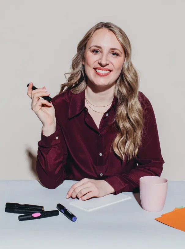 Melanie Bosse, lächelnd in dunkelroter Bluse an einem Tisch mit Stiften und Kaffeetasse