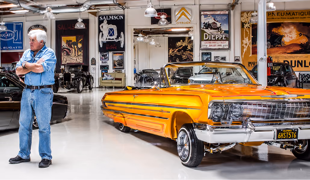 Man in blue denim outfit standing beside an orange classic convertible car inside a vintage car showroom.
