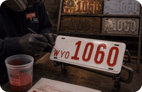 Person hand-painting red letters on a white Wyoming license plate, with old plates in the background.