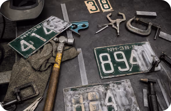 Worktable with weathered license plates, hammer, clamps, and various metalworking tools scattered on surface.
