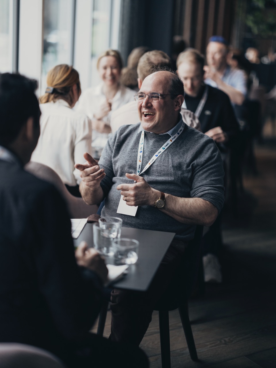 Mann med briller og grå genser ler og gestikulerer mens han snakker med en annen person på en kafé eller konferanse, med flere mennesker i bakgrunnen.