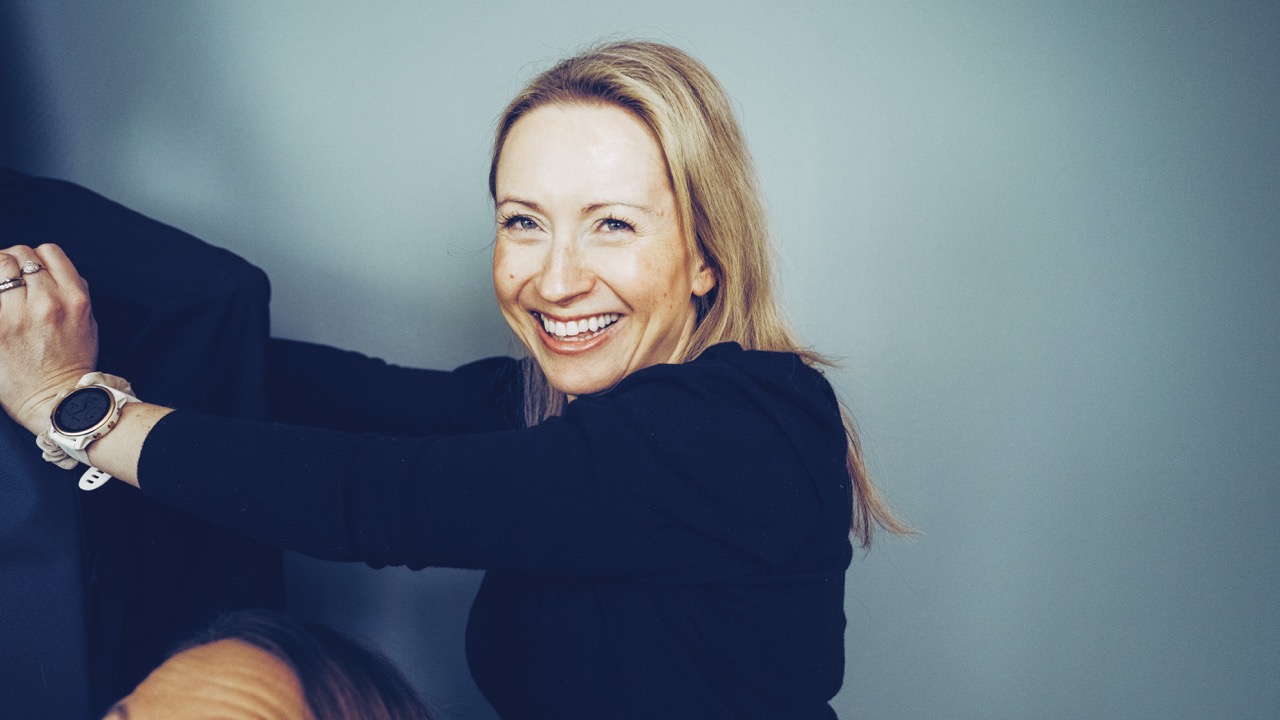 Smilende kvinne med blondt hår i svart genser foran en blå vegg.