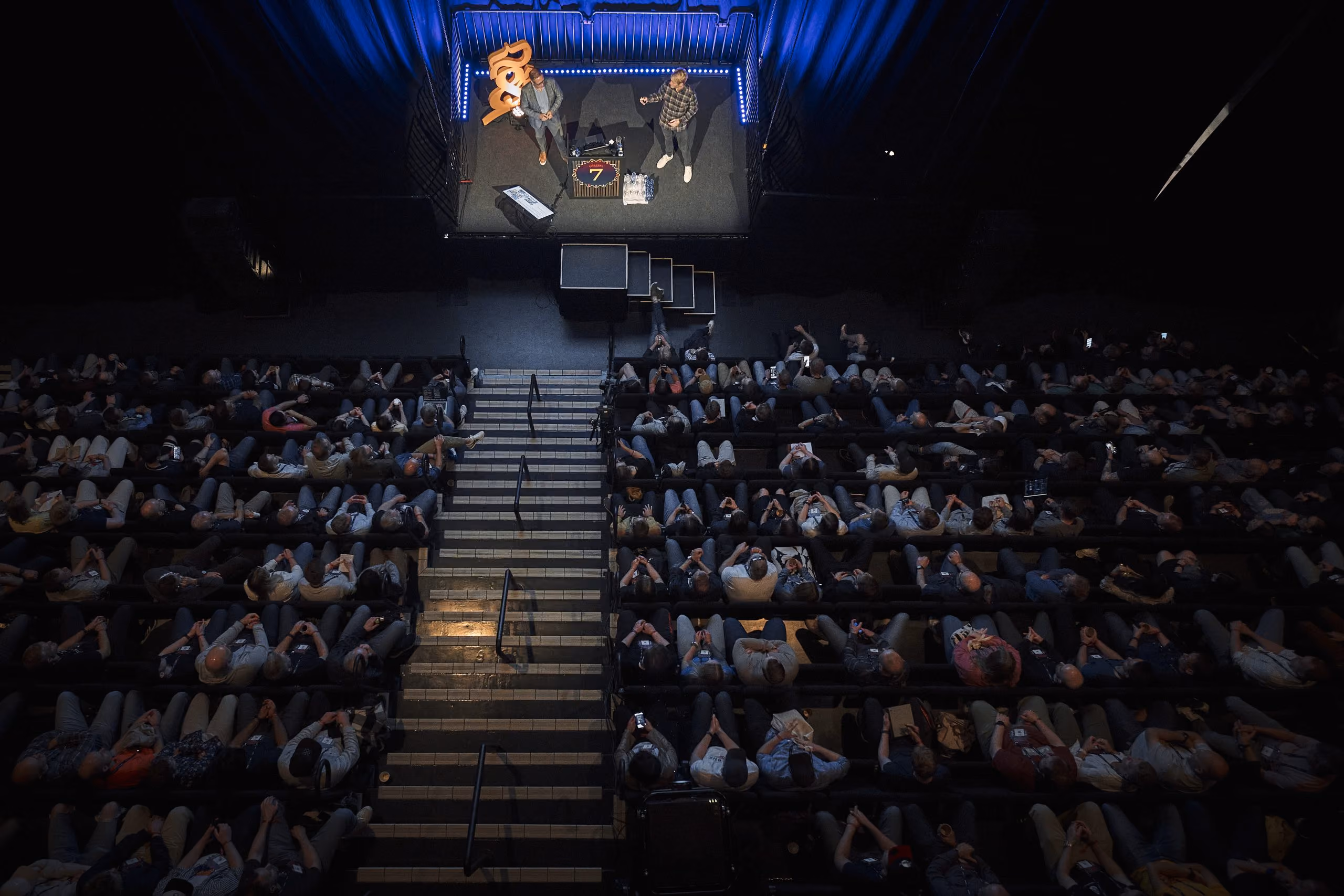Oversiktsbilde av et auditorium med to personer som holder et foredrag på en opplyst scene foran publikum.