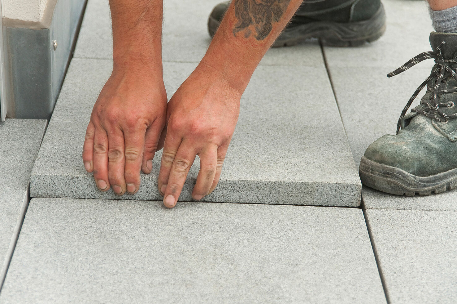 Zwei Hände positionieren eine graue Terrassenplatte exakt an der Fuge, Handwerker-Stiefel im Hintergrund