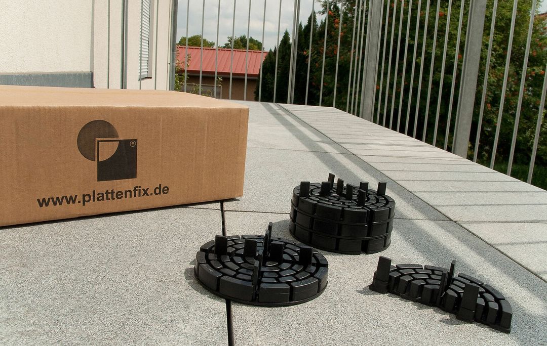 PLATTENFIX Versandkarton mit Firmenlogo neben montierten Stelzlagern auf einer Terrasse