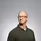 Smiling elderly man with glasses, wearing a dark sweater against a plain gray background.
