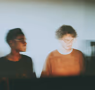 Blurred image of two people, one darker-skinned and one lighter-skinned, standing side by side indoors.