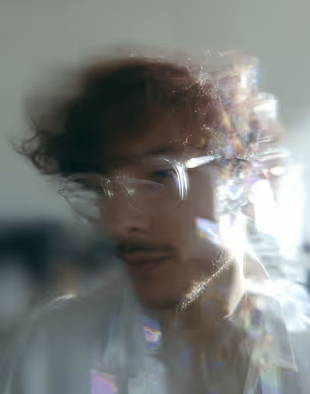 Blurred close-up of a man with glasses and dark hair amid light reflections and distortions.
