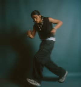Person in black clothing and sneakers dancing against a plain dark teal background.