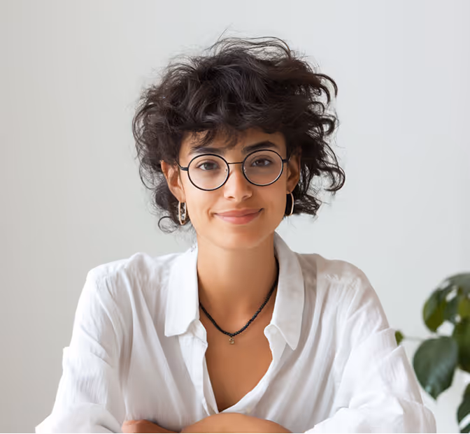Smiling woman with curly hair and glasses wearing a white shirt in a bright room with a plant.