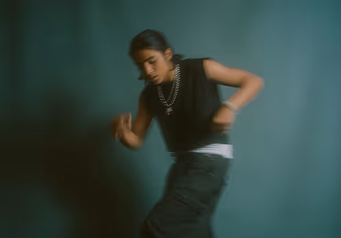 Blurred image of a person dancing wearing a black sleeveless top and dark pants against a plain backdrop.