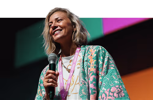 Smiling woman holding microphone, wearing a colorful floral jacket and a purple lanyard on stage.