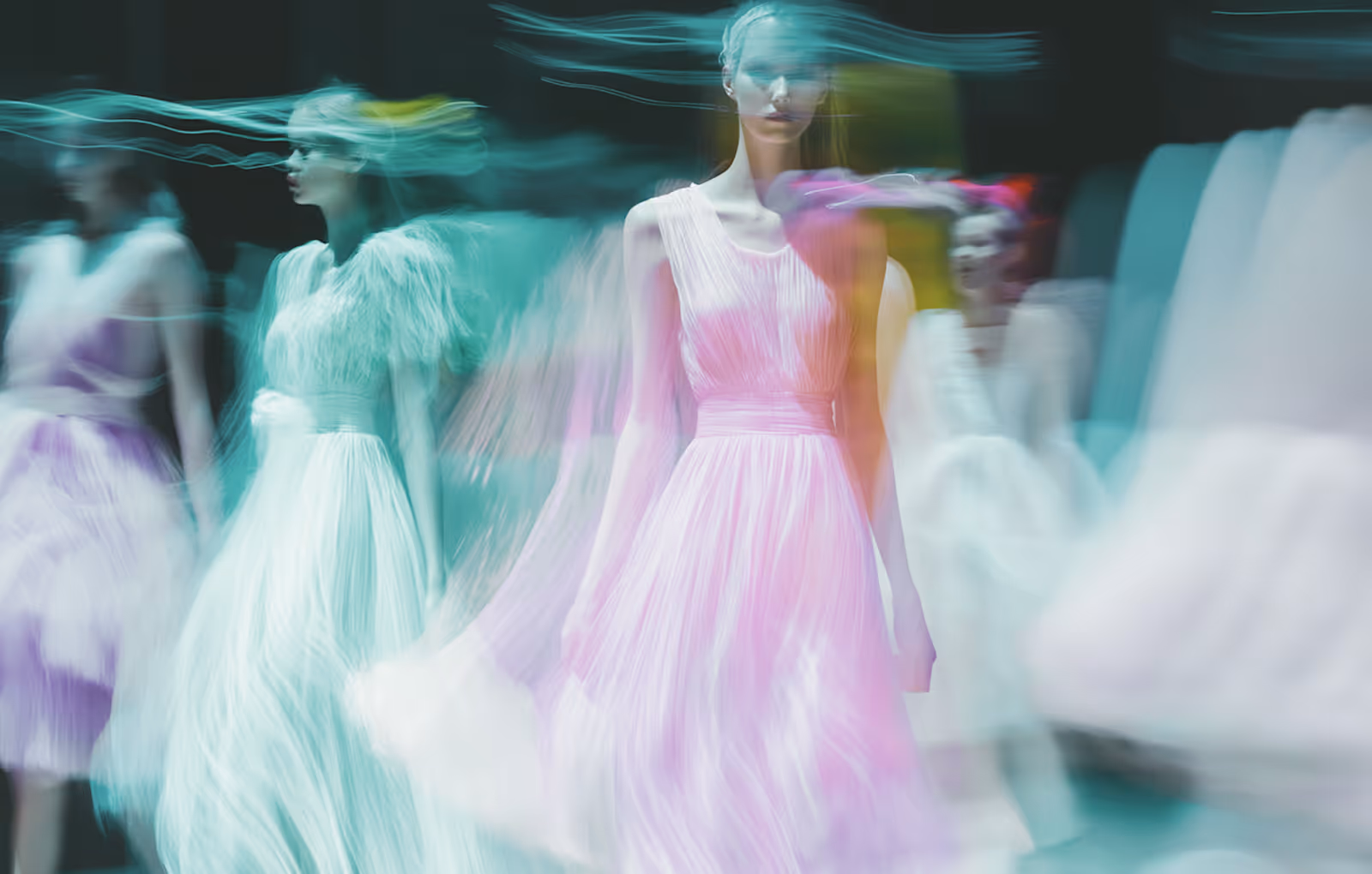 Motion-blurred image of models wearing flowing pastel dresses on a dark runway.