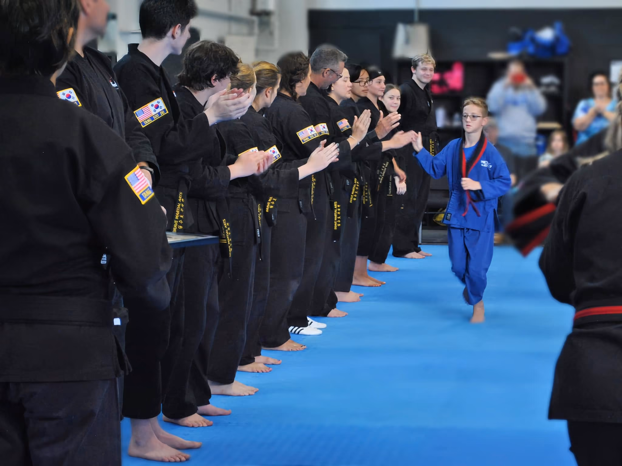 A young martial artist in a blue uniform walks confidently past applauding peers in black uniforms on a blue mat.