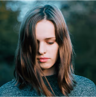 Young woman with long brown hair looking down, wearing a blue knit sweater, with a blurred green natural background.