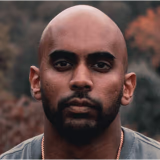 Close-up portrait of a bald man with a dark beard and mustache, looking directly at the camera with a neutral expression, outdoors with blurred natural background.