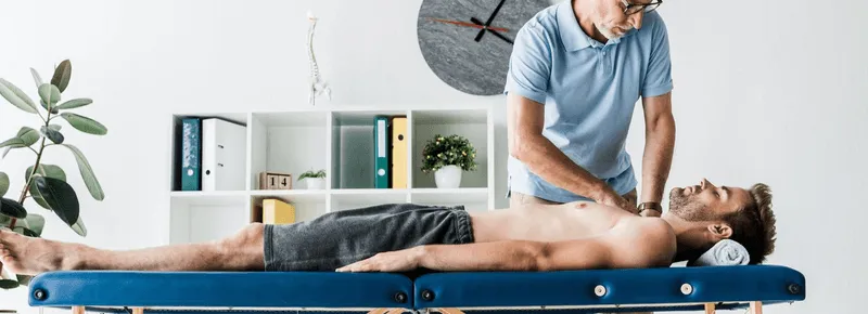 Therapist in glasses performing chest therapy on a shirtless man lying on a blue massage table in a bright treatment room with a plant and shelves in the background.
